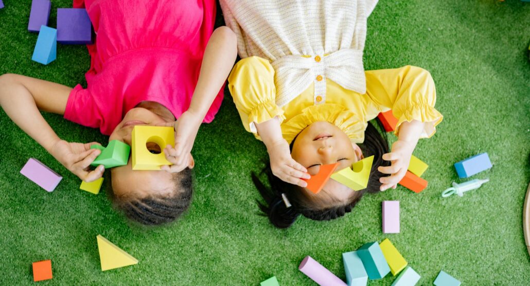 Two little girls lying down playing with wooden blocks.